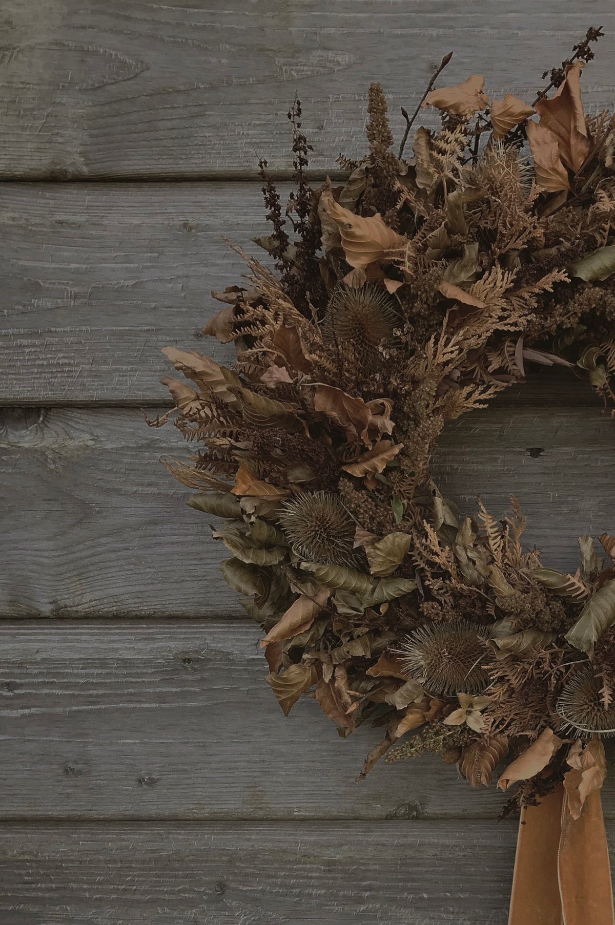 A dried floral wreath made of brown leaves, thistles, and ferns, hanging on a weathered gray wooden wall.