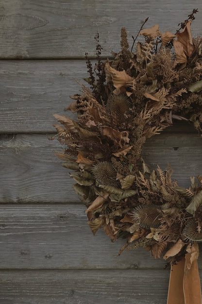 A dried floral wreath made of brown leaves, thistles, and ferns, hanging on a weathered gray wooden wall.