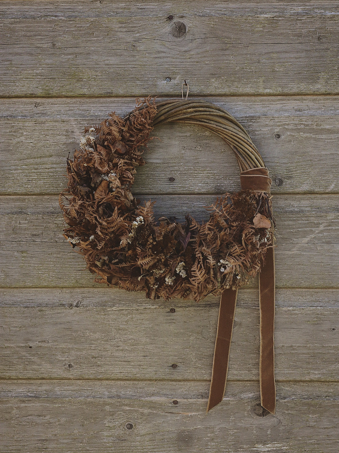 A rustic brown wreath made from dried ferns and small white flowers, hanging on a weathered wooden plank wall with a brown ribbon bow and two hanging tails.