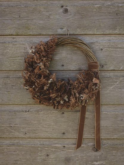 A rustic brown wreath made from dried ferns and small white flowers, hanging on a weathered wooden plank wall with a brown ribbon bow and two hanging tails.