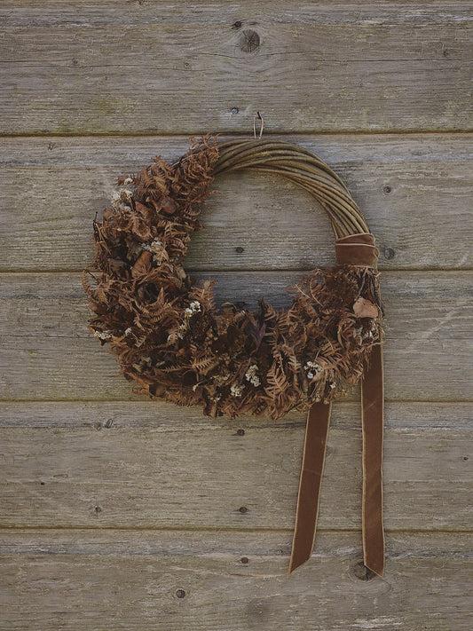 A rustic brown wreath made from dried ferns and small white flowers, hanging on a weathered wooden plank wall with a brown ribbon bow and two hanging tails.