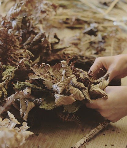 A close-up of hands arranging a dried botanical wreath made of brown leaves, ferns, and pinecones on a wooden surface.