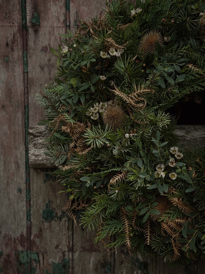 A rustic green wreath made of pine branches, dried ferns, thistle-like pods, and small white flowers, hanging against weathered wooden doors.