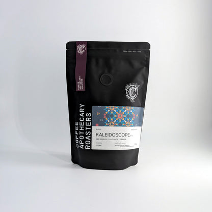 A black stand-up pouch of Coffee Roastery Kaleidoscope blend coffee beans, featuring a colorful geometric label and a purple vertical stripe, set against a clean white studio background.