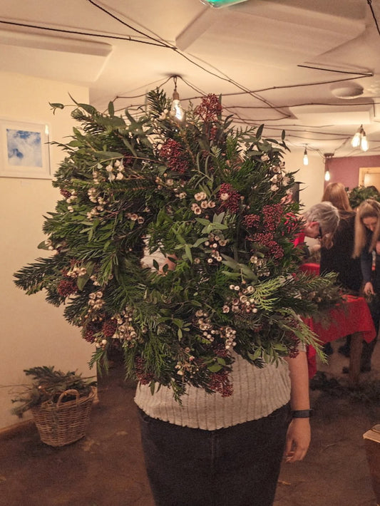 A large, lush Christmas wreath made of green pine and fir branches, decorated with red berries and small white flowers, held by a person in a ribbed cream sweater in an indoor event space.